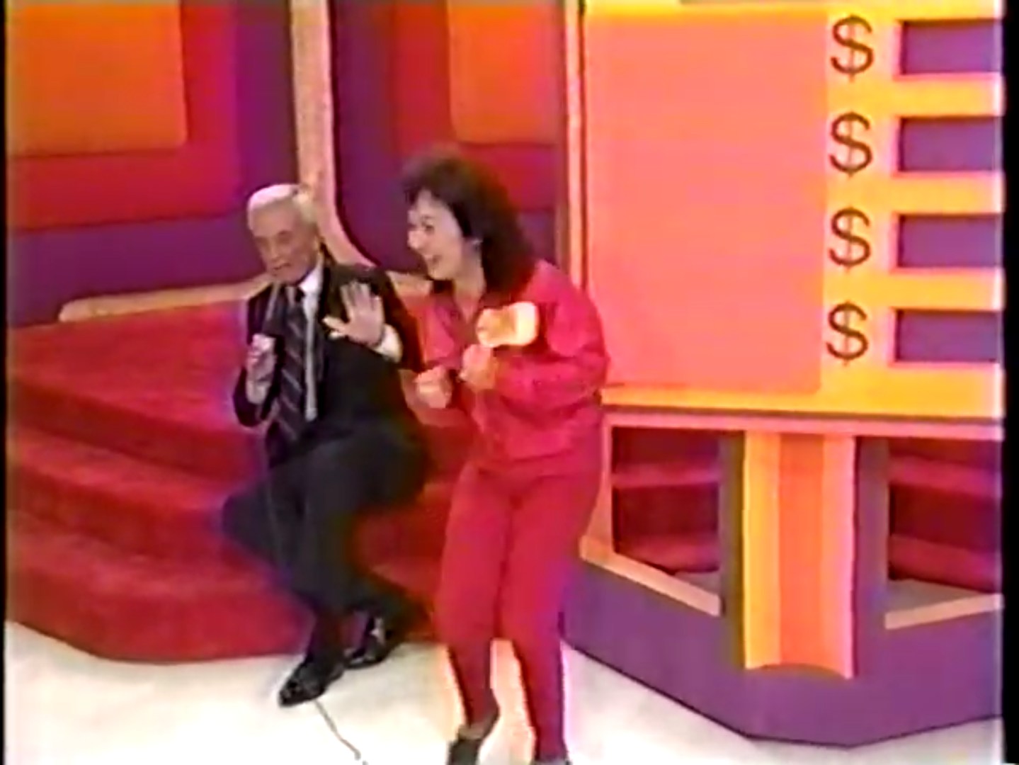 A contestant in a red outfit celebrates excitedly next to a host on stage, with a colorful game board displaying dollar signs in the background.