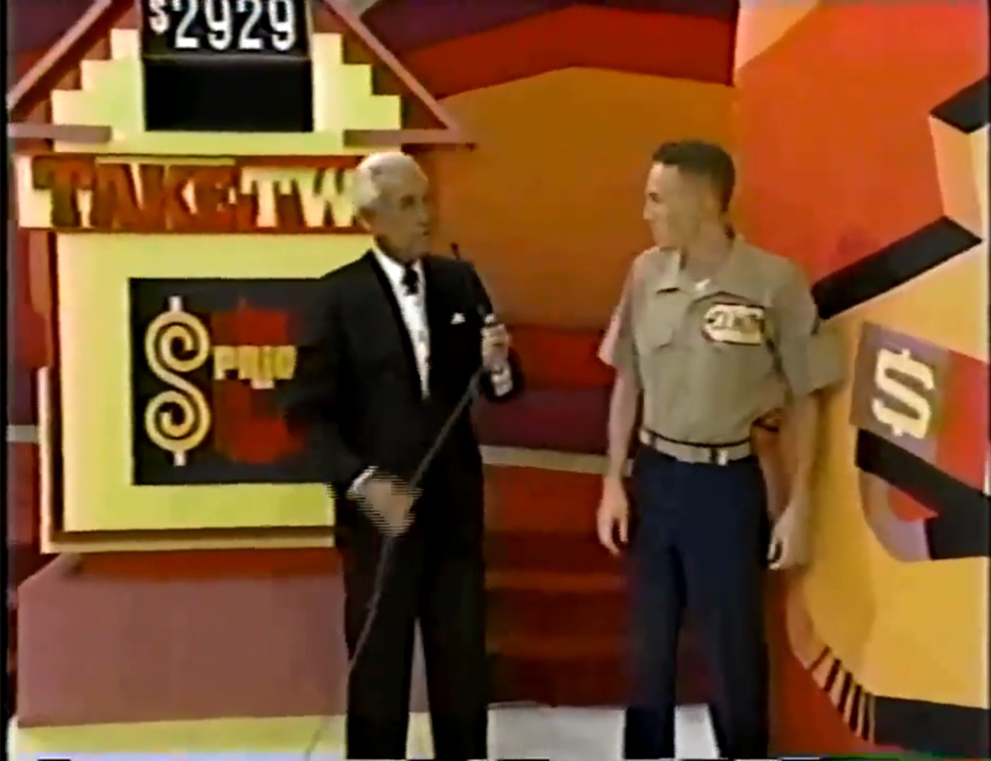 A game show host stands next to a contestant during the Take Two game on The Price is Right, with a colorful set and a sign displaying the target price.