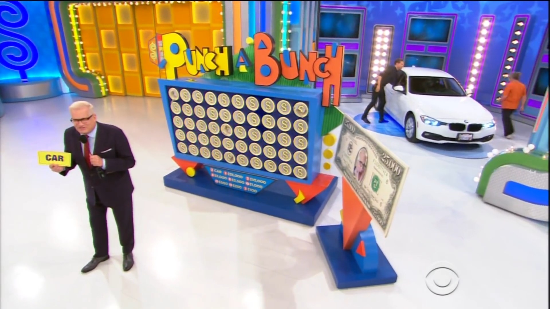 A game show host stands in front of a colorful Punch a Bunch board while holding a card labeled 'CAR,' with a white BMW visible in the background and a ,000 bill displayed.