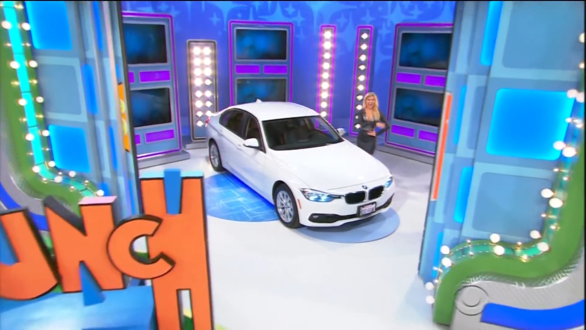 A white BMW car is showcased on stage at The Price is Right, with colorful lights and a host standing nearby.