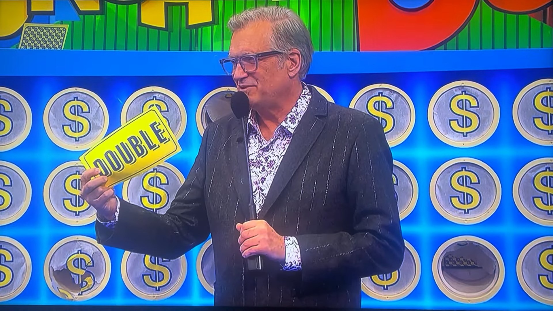 A host from The Price is Right holds up a yellow 'DOUBLE' slip while standing in front of the punchboard featuring money symbols.