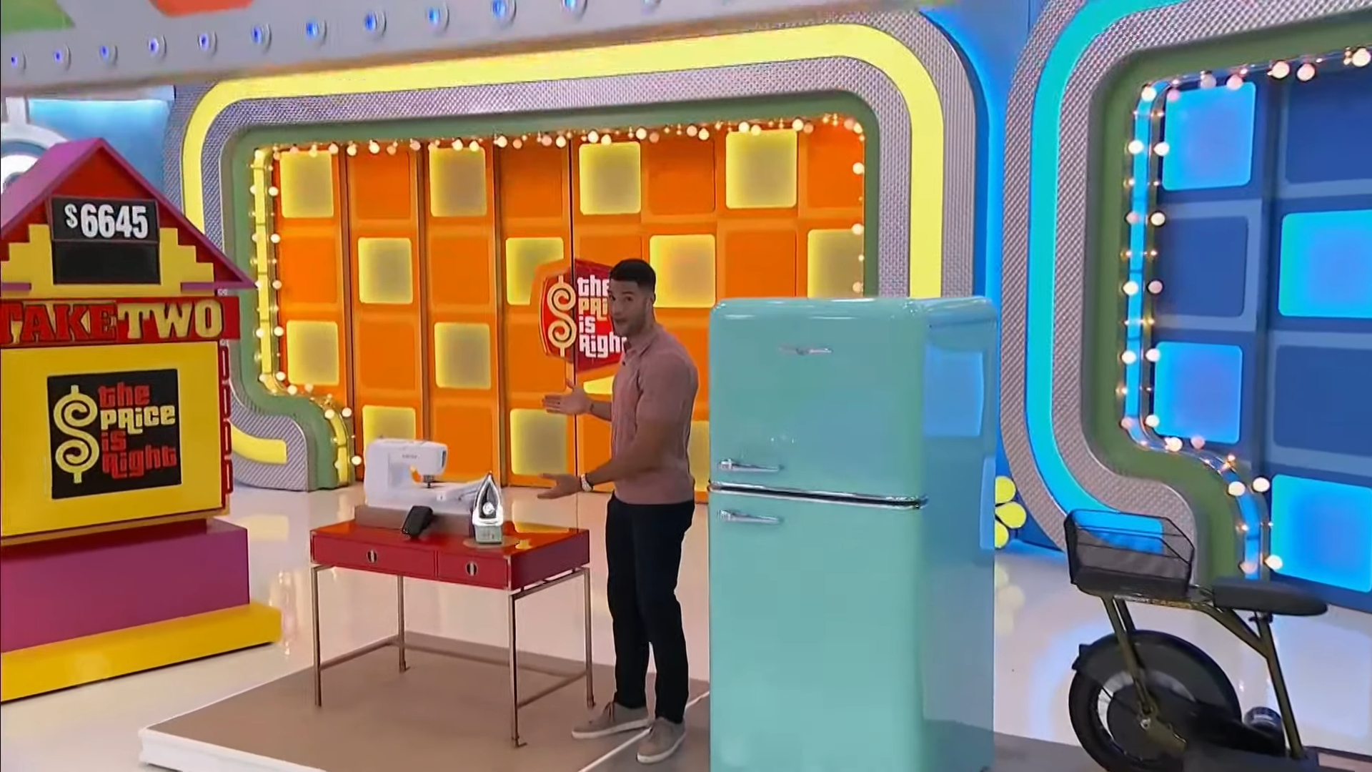 A contestant stands beside the 'Take Two' game board on 'The Price is Right' set, with a variety of prizes displayed, including a retro refrigerator, a sewing machine, an iron, and a bicycle.