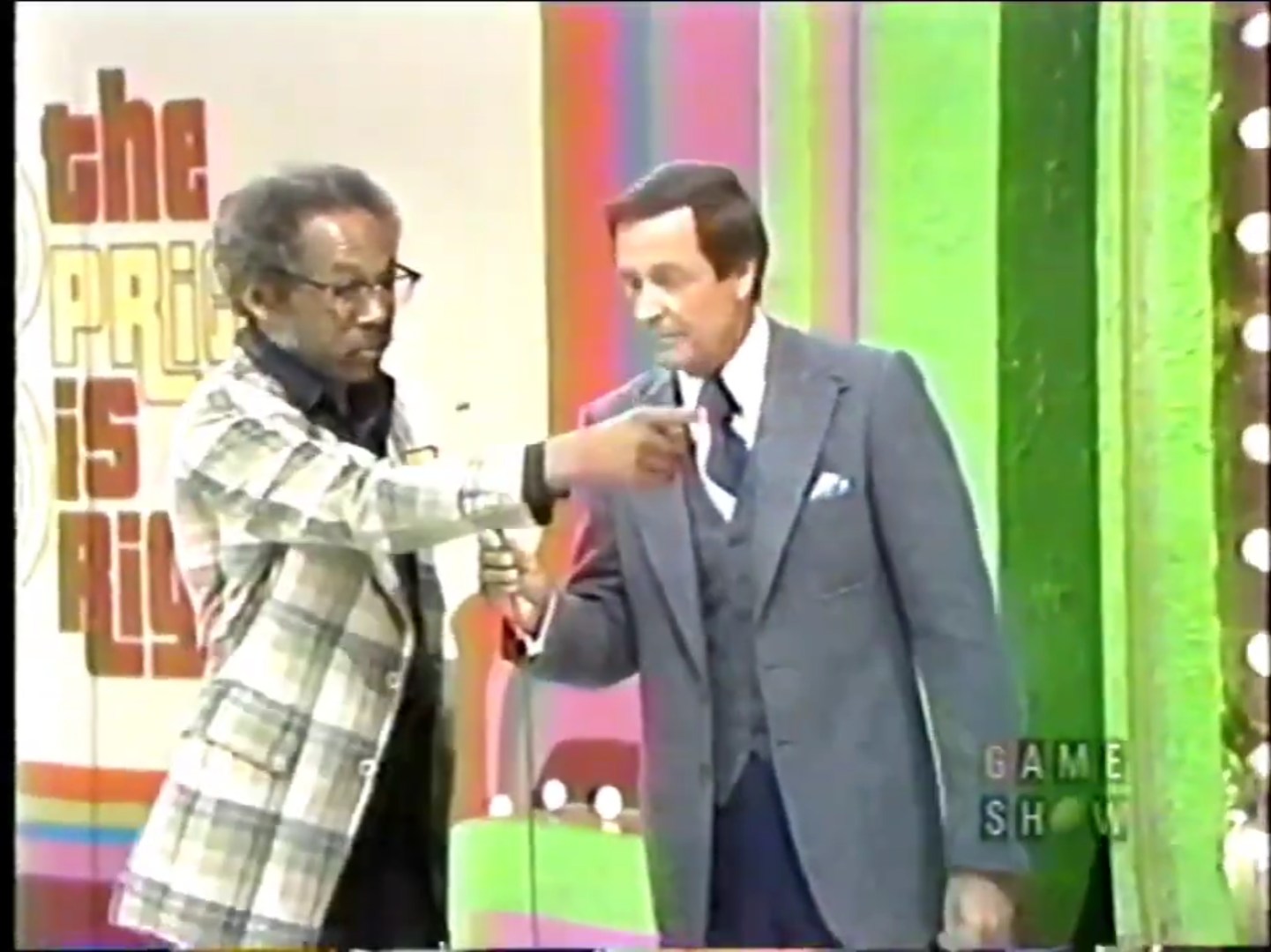 An African American contestant in a plaid blazer interacts with the host on a game show stage decorated in vibrant colors.