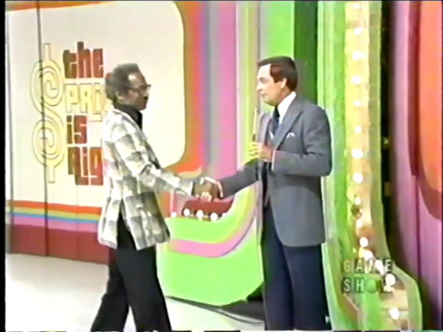 A contestant shakes hands with the host on the set of The Price is Right game show, featuring colorful backgrounds and a prominent logo.