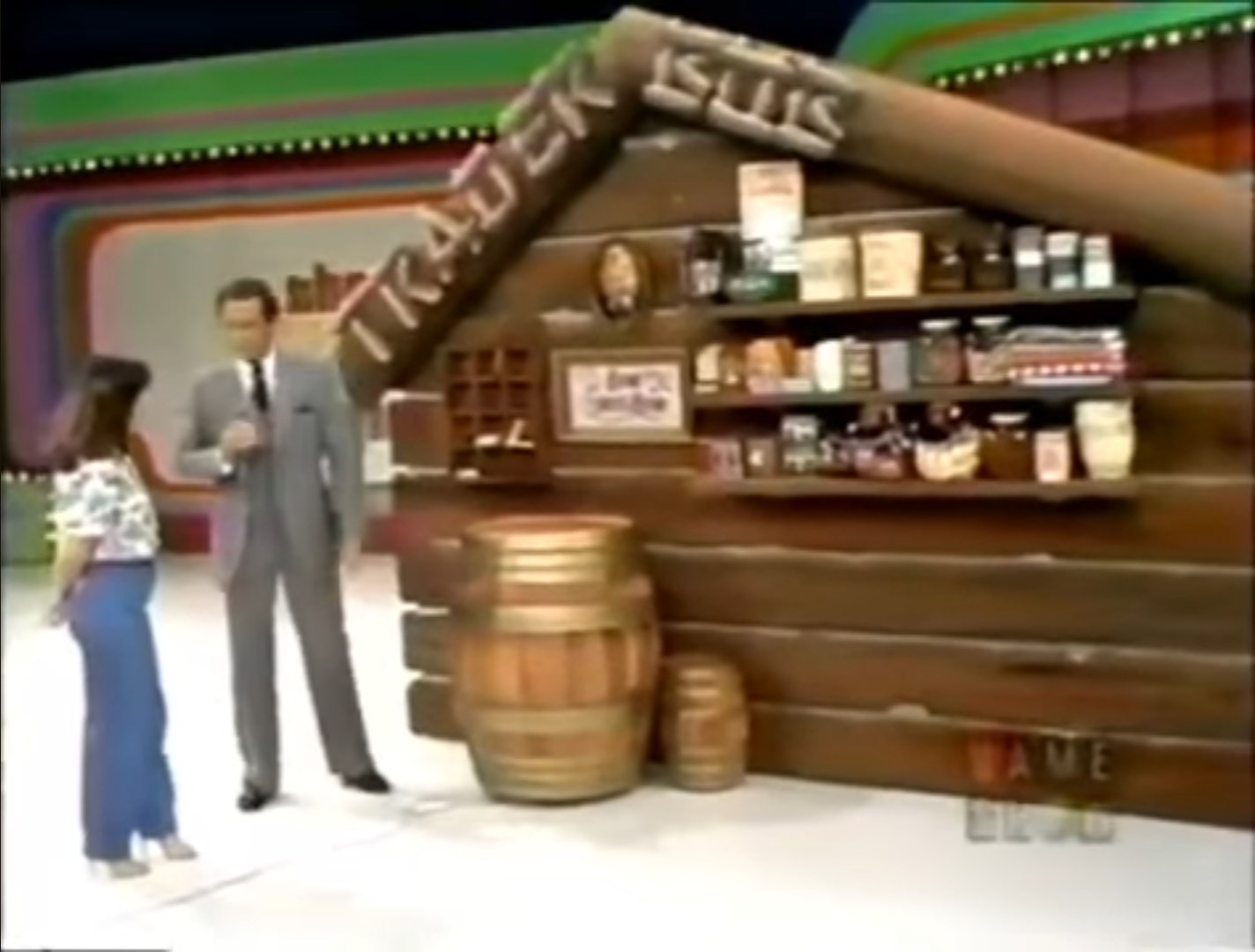 A rustic cabin-themed set for the game 'Trader Bob' featuring shelves with prizes and a contestant interacting with Bob Barker.