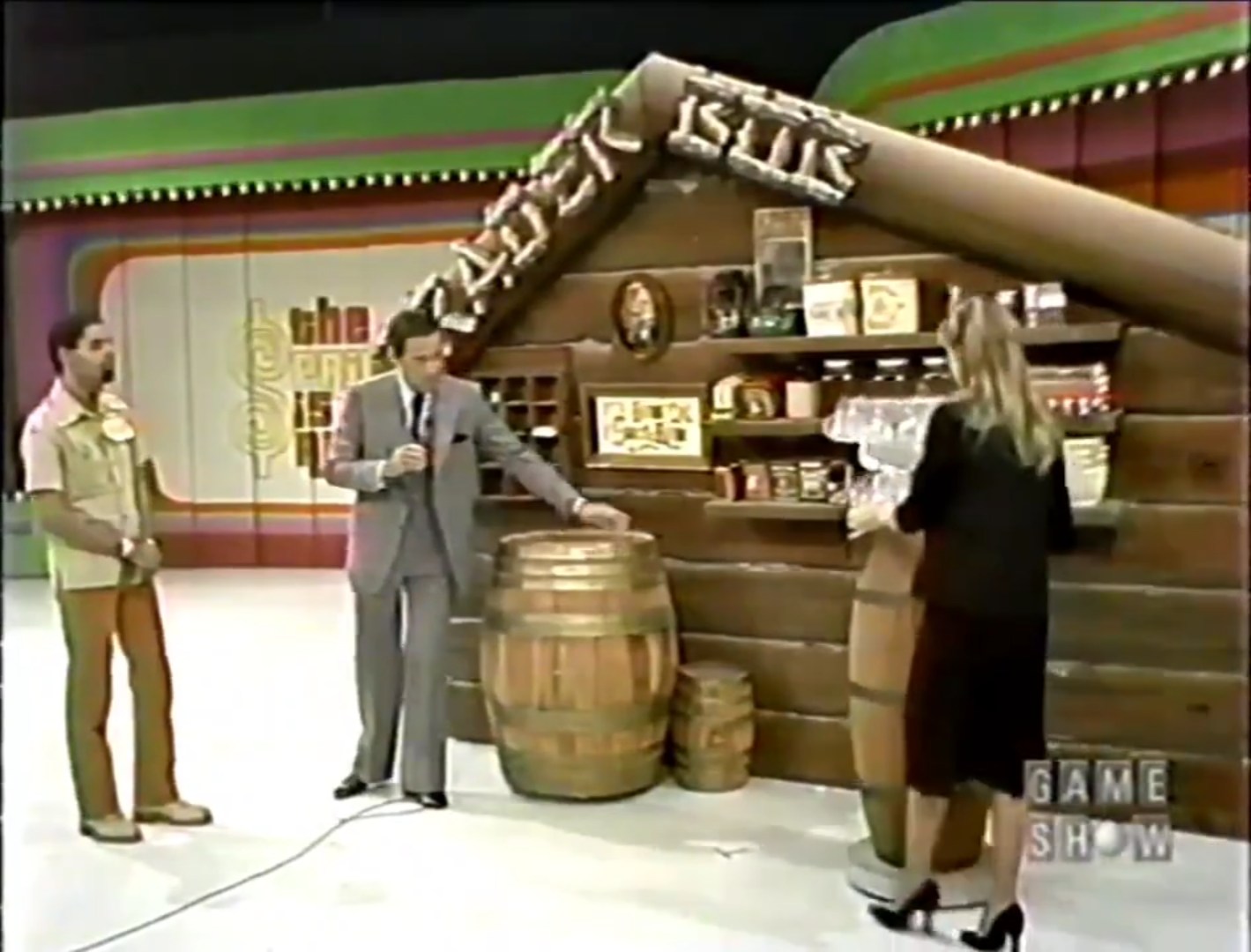 A game show scene featuring Trader Bob's rustic cabin-themed set, with Bob Barker speaking into a microphone and a female contestant interacting with a display or prize.
