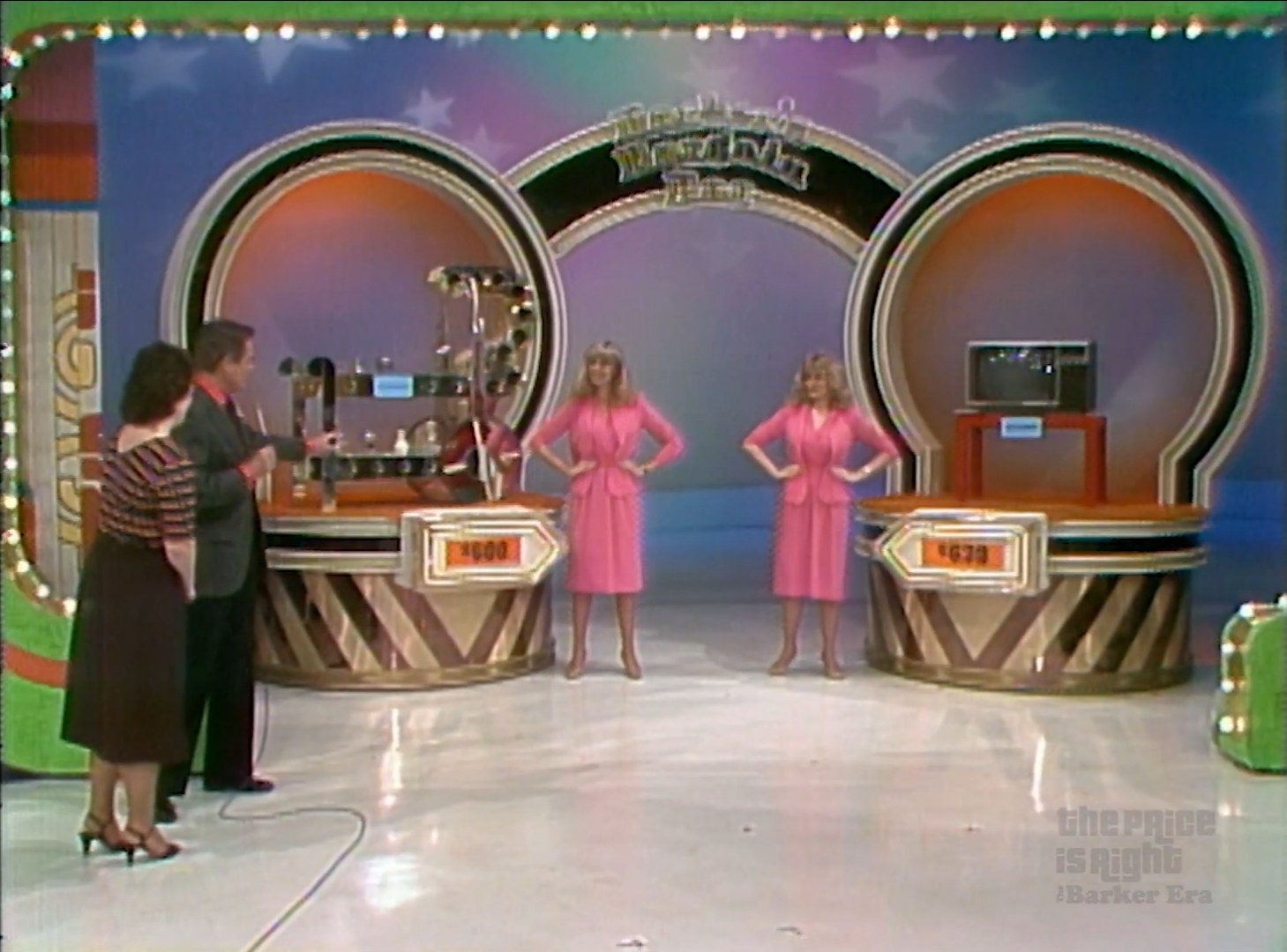 A scene from the Bargain Game on The Price is Right featuring a host presenting two prizes with two models in pink dresses, one prize costing 0, the other 8, set against a colorful stage backdrop.