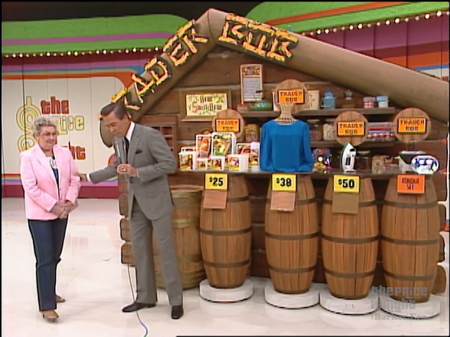 A game show set featuring Trader Bob with wooden barrels displaying various prizes, including a blue shirt and an iron, alongside a contestant and the host.