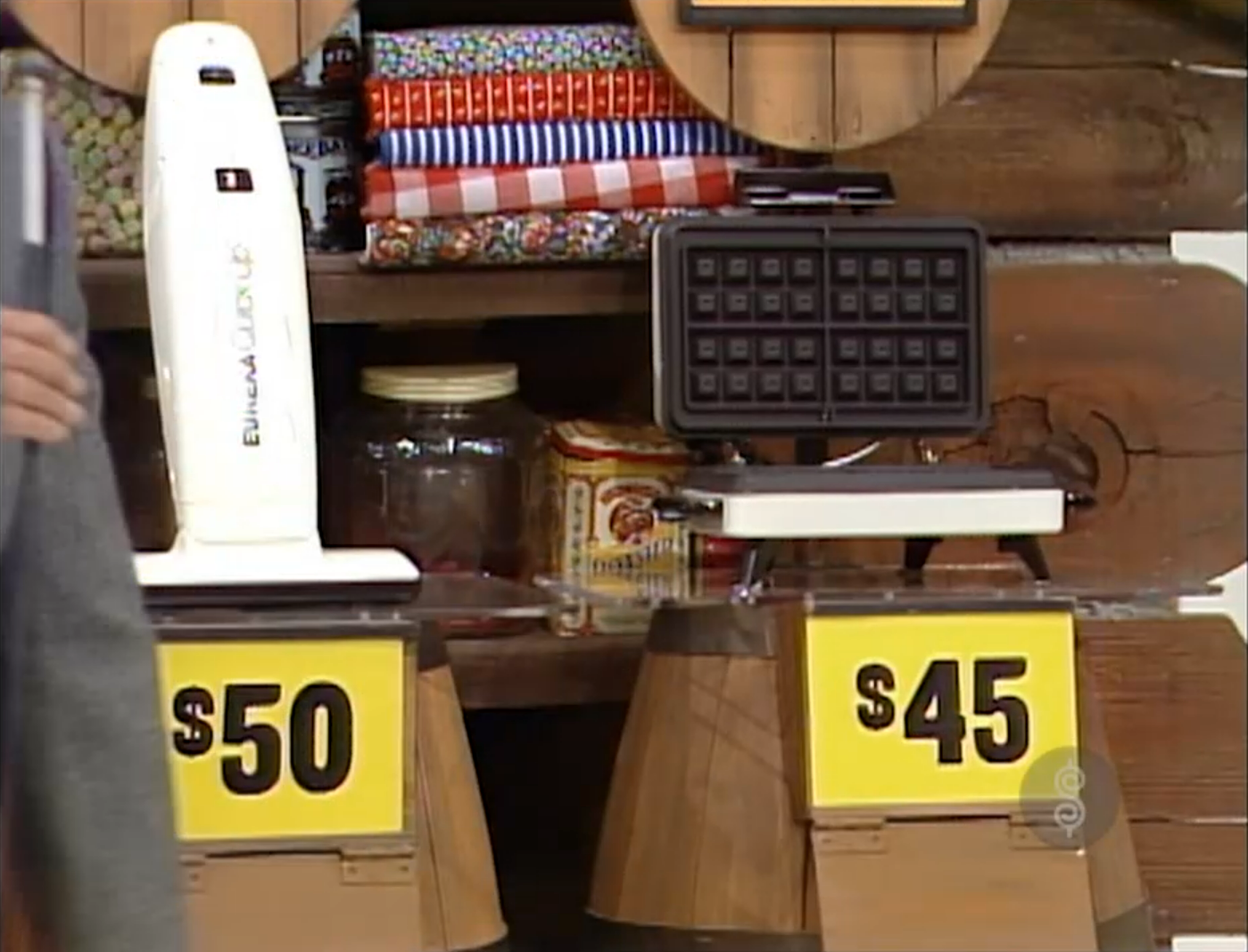 Two prize items: a white appliance priced at  and a black waffle maker priced at , displayed on wooden pedestals in a rustic setting.