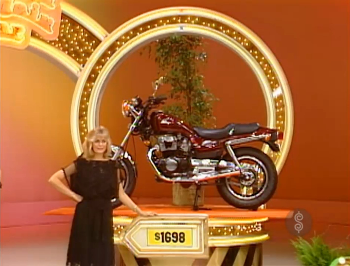 A contestant stands next to a motorcycle showcased on a pedestal with a price tag of 98, set against a colorful backdrop.