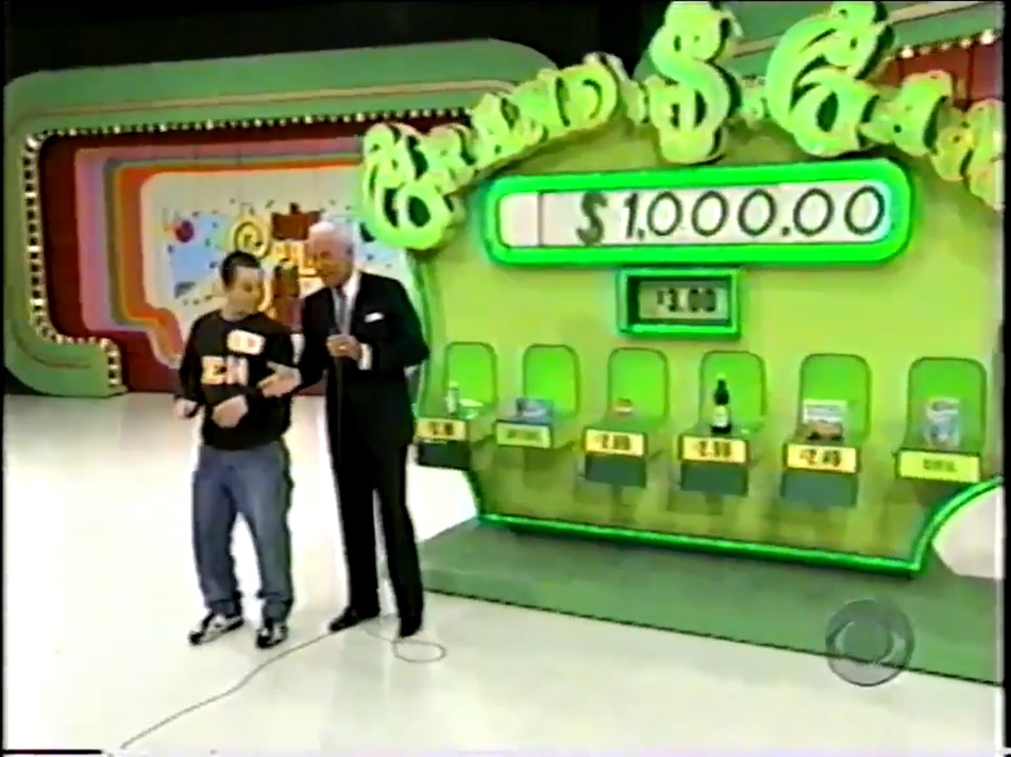 Contestant and host interact in the Grand Game setup, with a prize board displaying ,000 and prices of grocery items below.