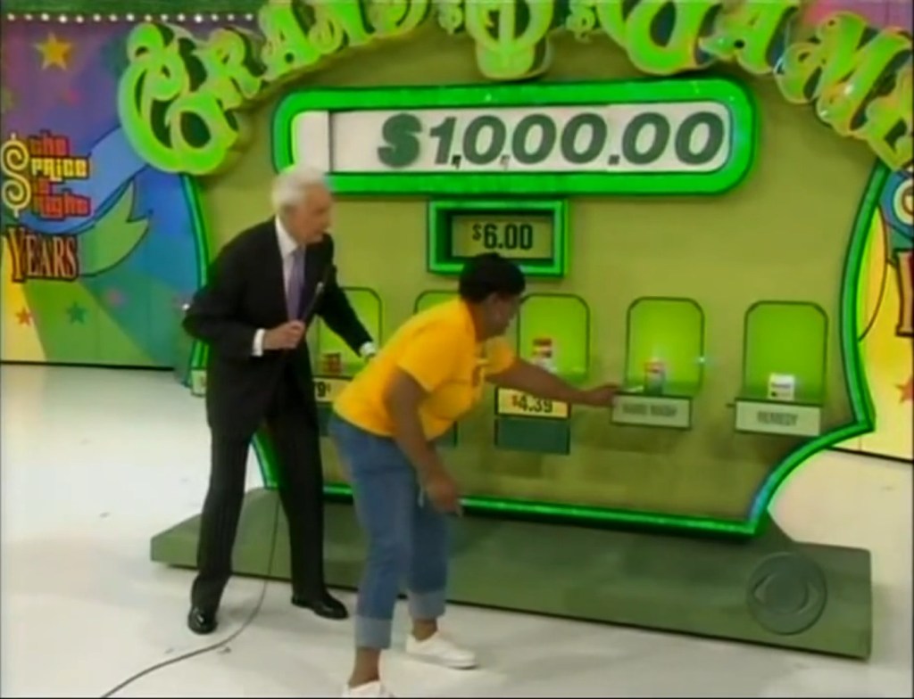 Contestant selecting a grocery item on The Price is Right's Grand Game, with host Bob Barker nearby and the game's prize displayed behind.