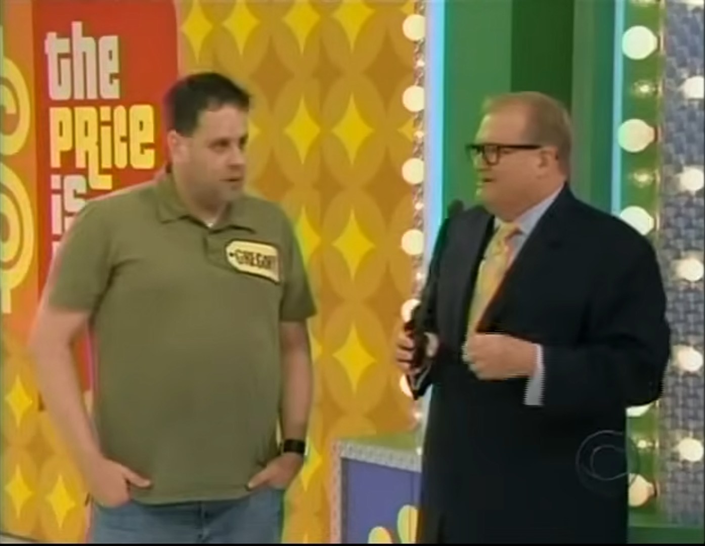Contestant Greg stands on stage during the Bargain Game segment of The Price is Right, with host Drew Carey beside him against a colorful game show backdrop.