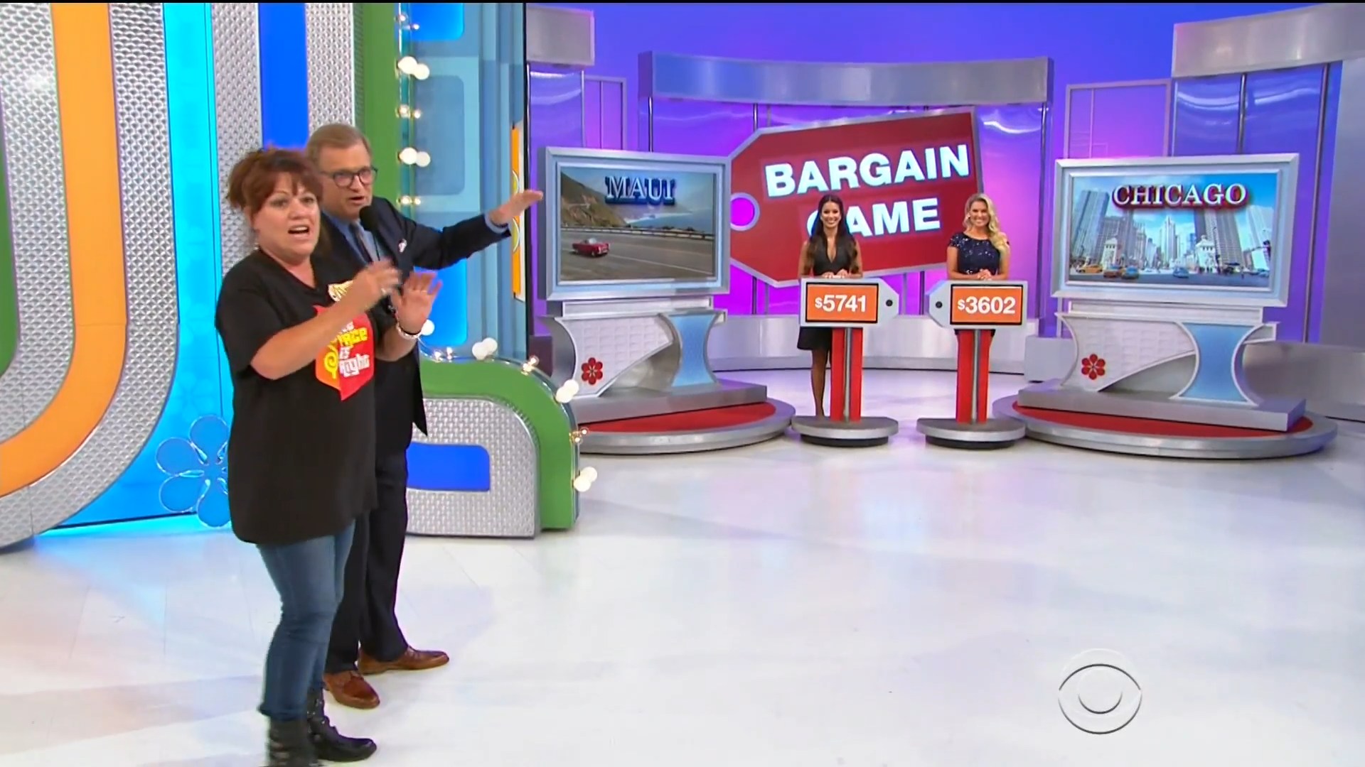 A scene from the Bargain Game on The Price is Right, featuring a host and a female contestant excitedly discussing prizes, with two large display screens showcasing locations and their associated prices.