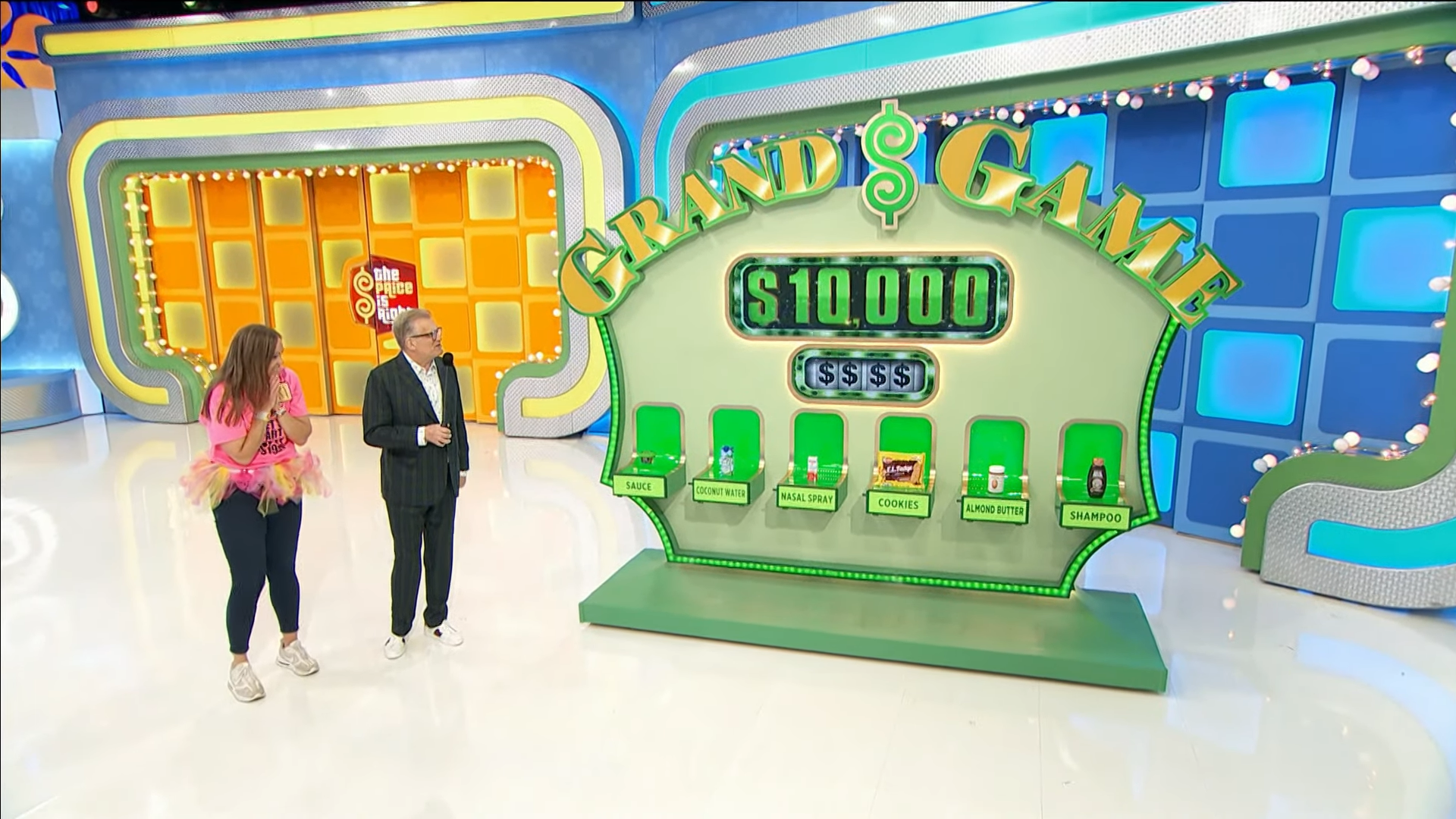 A contestant, wearing a pink shirt and tutu, reacts excitedly next to the host during the Grand Game on The Price is Right. The game board displays a target prize of $10,000 and six grocery items including sauce, coconut water, nasal spray, cookies, almond butter, and shampoo.