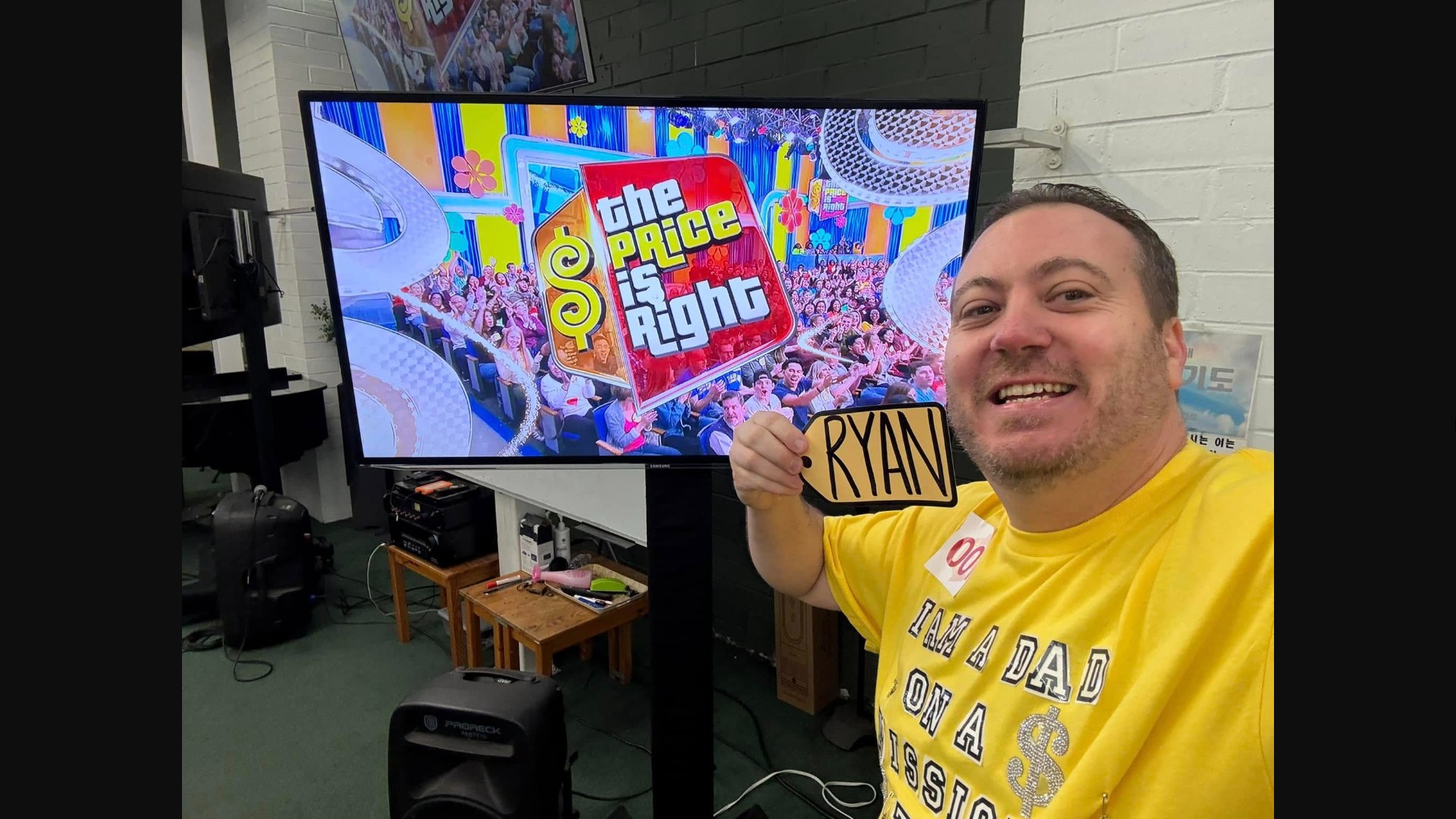 A Price is Right contestant named Ryan in a yellow shirt proudly shows off his name tag in the waiting area at The Price is Right.