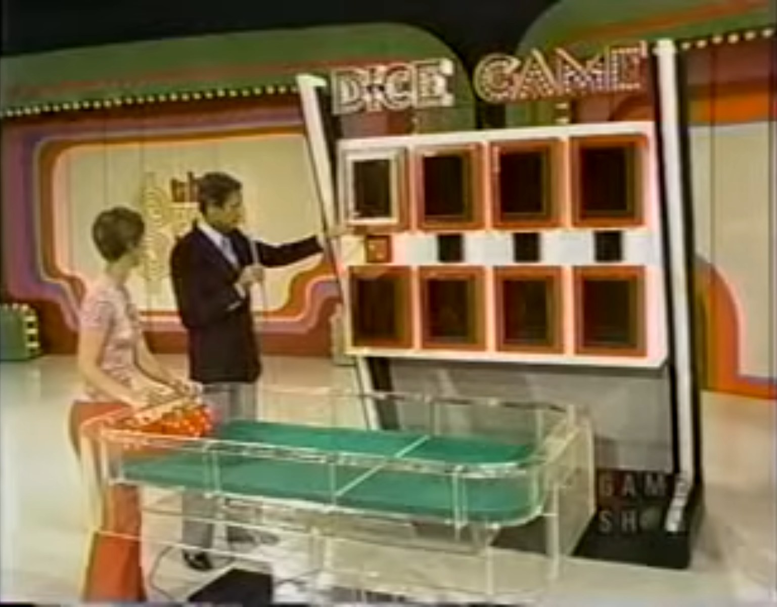 A contestant and host at the Dice Game setup, featuring oversized dice and a game board with hidden numbers.