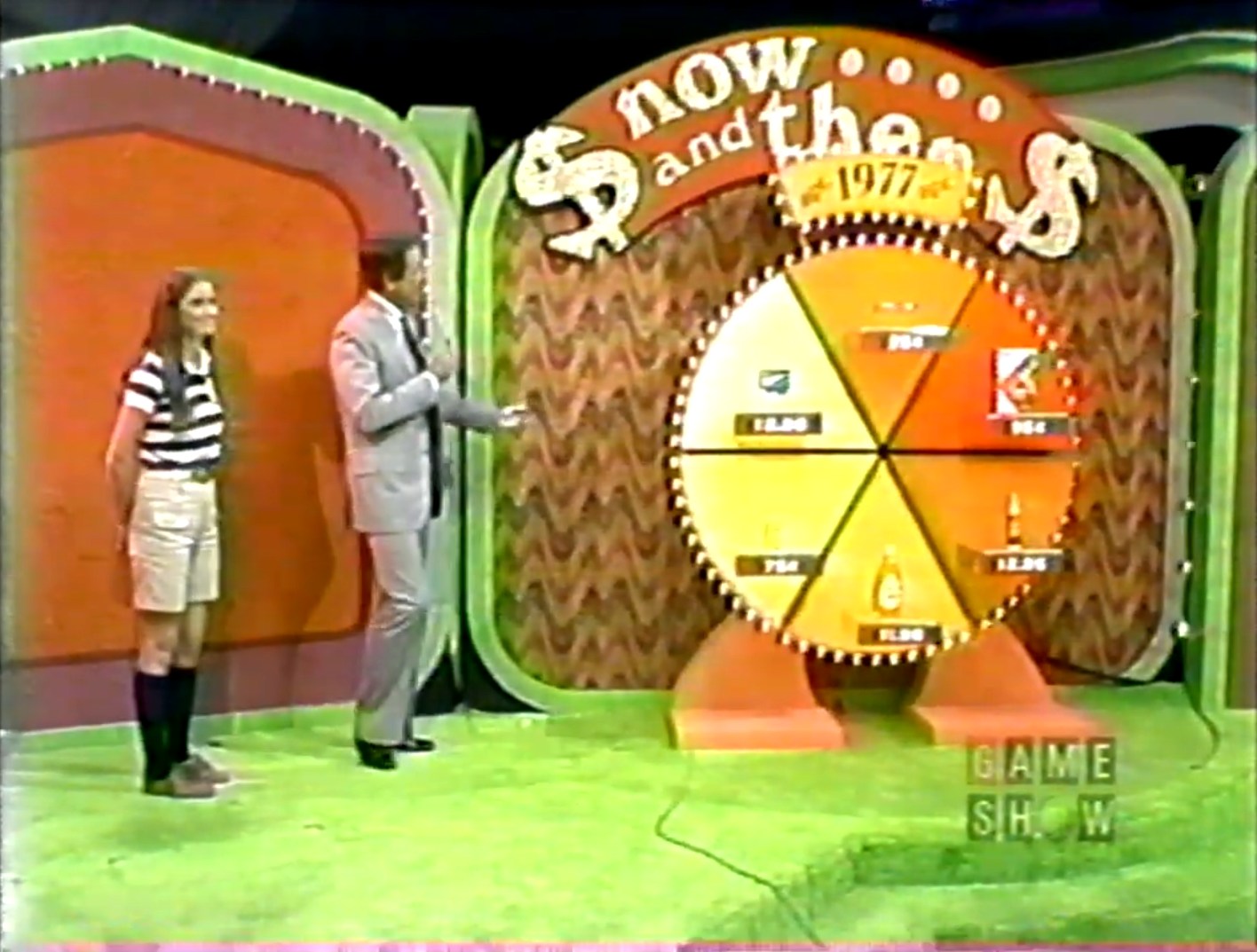 A game show scene featuring a circular board labeled 'Now and Then 1977' with six grocery product prices displayed. A contestant stands on the left, while the host points to the board.