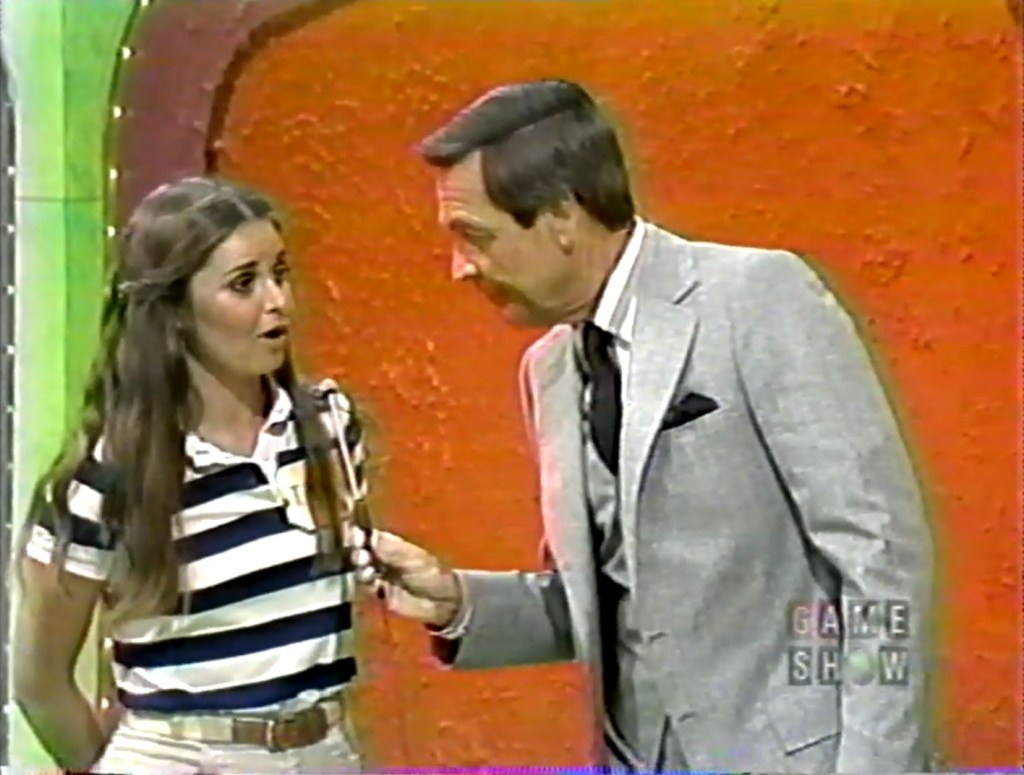 A Price is Right contestant, looking surprised, speaks to the host while holding a microphone, with a colorful background.