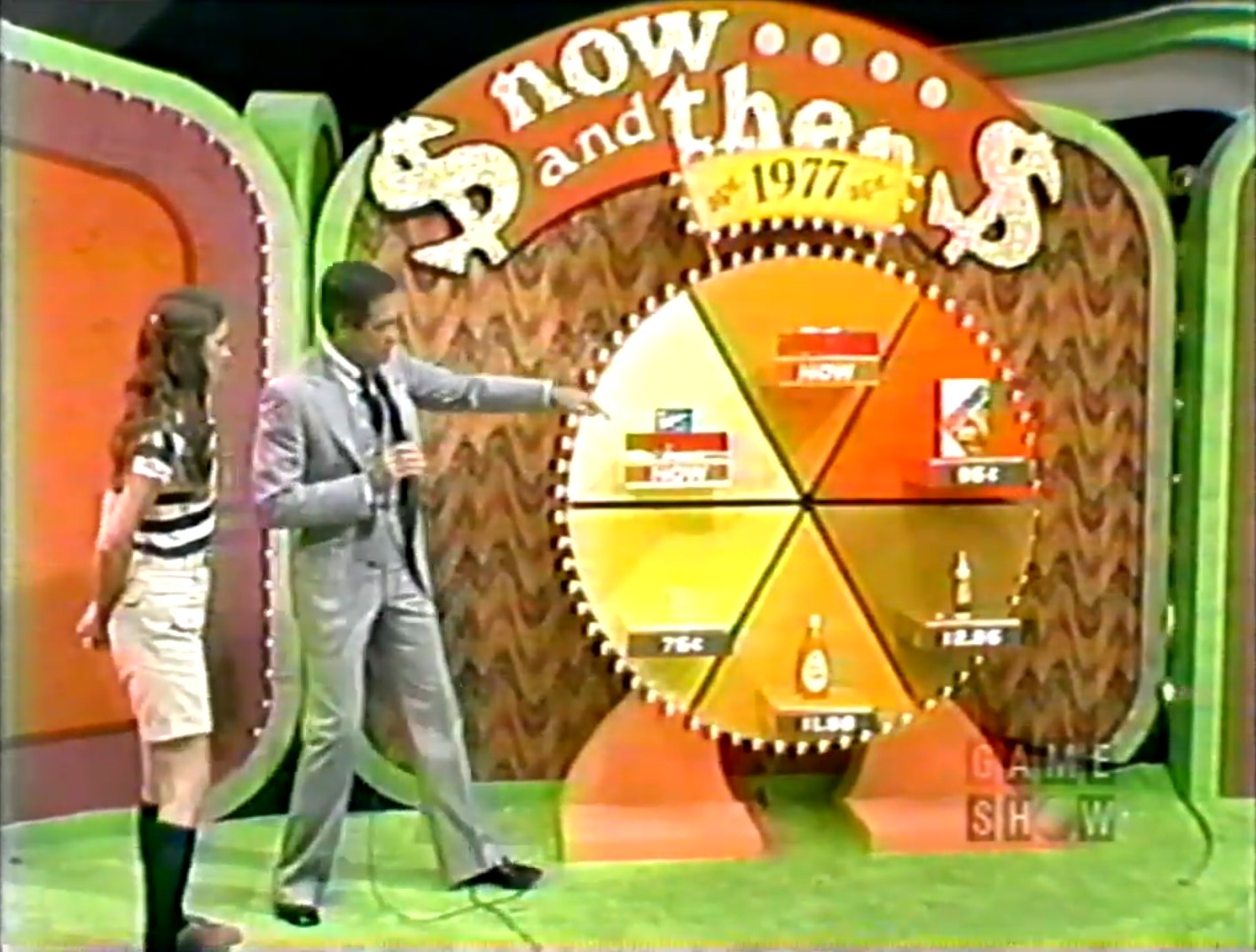 A game show scene featuring a host pointing at a circular board labeled 'Now and Then' with prices for various grocery products, while a female contestant stands beside him.