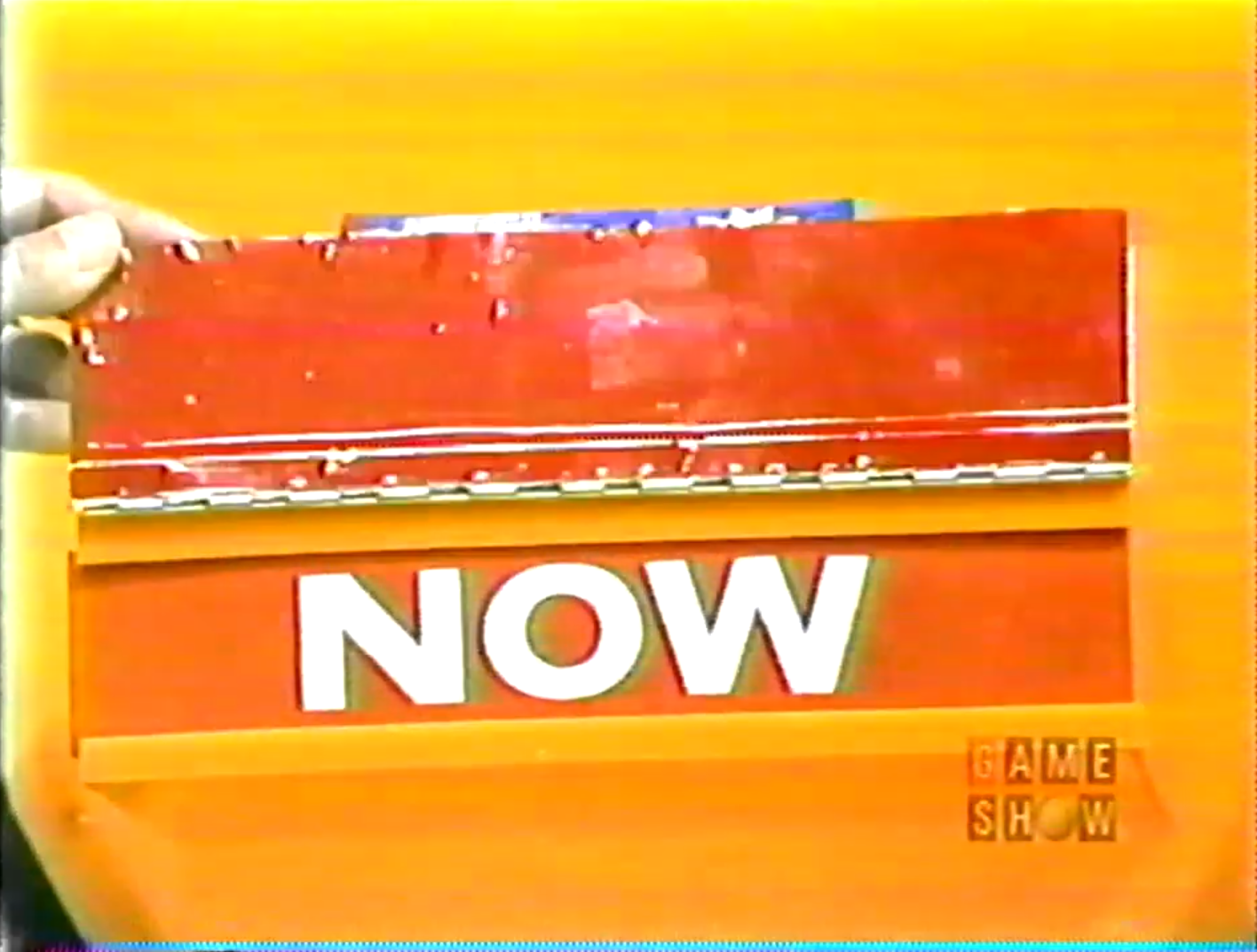 A close-up of a vibrant red sign displaying the word 'NOW' in large white letters against an orange background, indicating the current pricing game in 'Now or Then'.