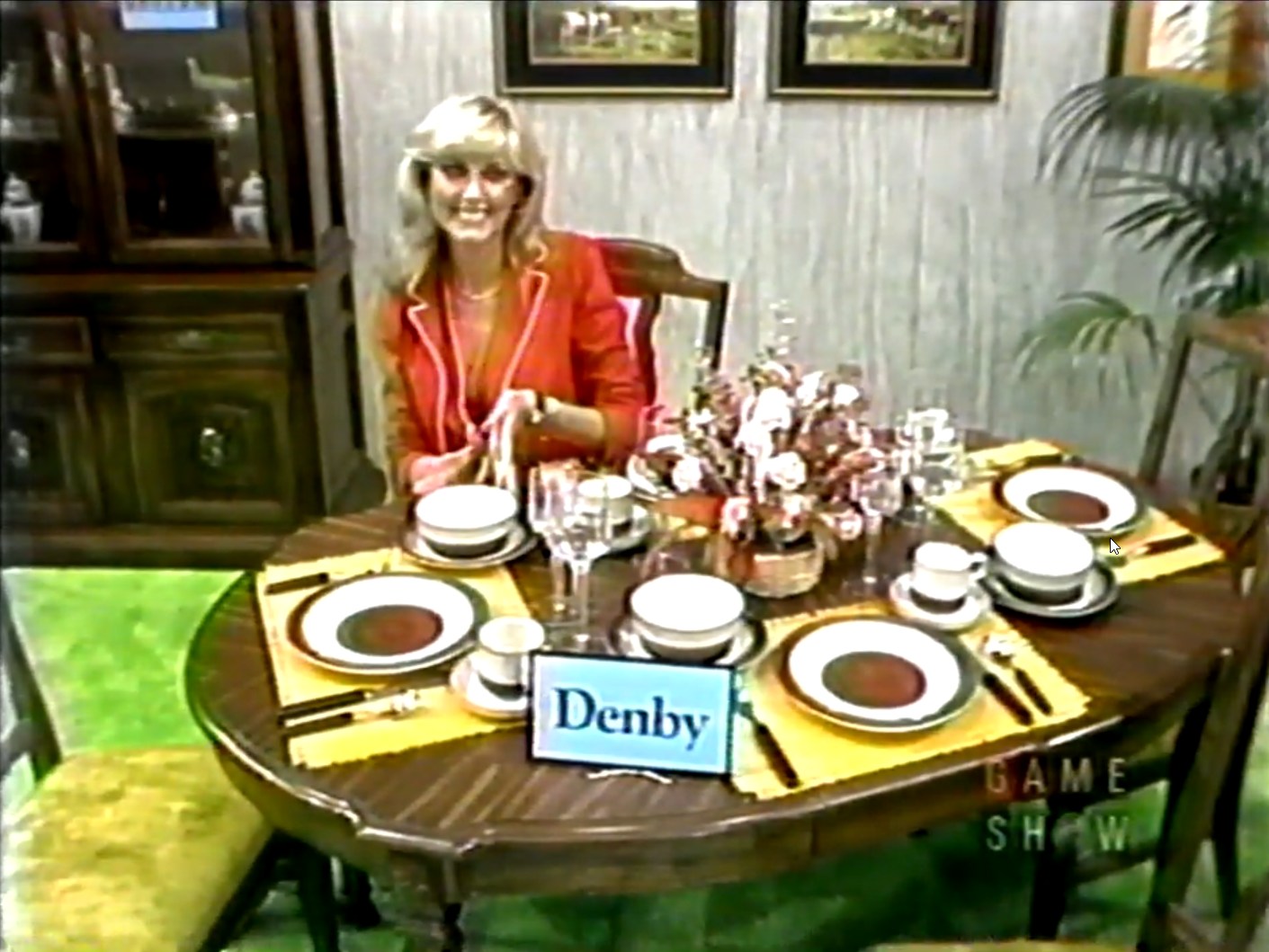 A smiling woman sits at a dining table set with plates, flatware, and glasses, showcasing a prize labeled 'Denby' in front of her.