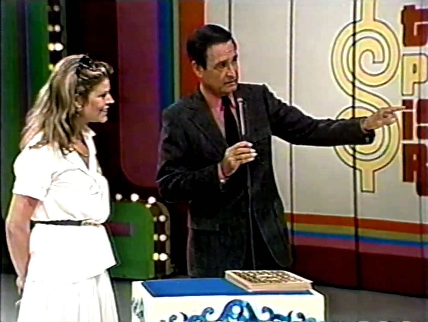 A contestant and the host of 'The Price is Right' are engaged in a pricing game. Bob Barker is pointing while speaking to the contestant, who is smiling. The colorful game show set background is visible. An oversized deck of cards in the foreground.