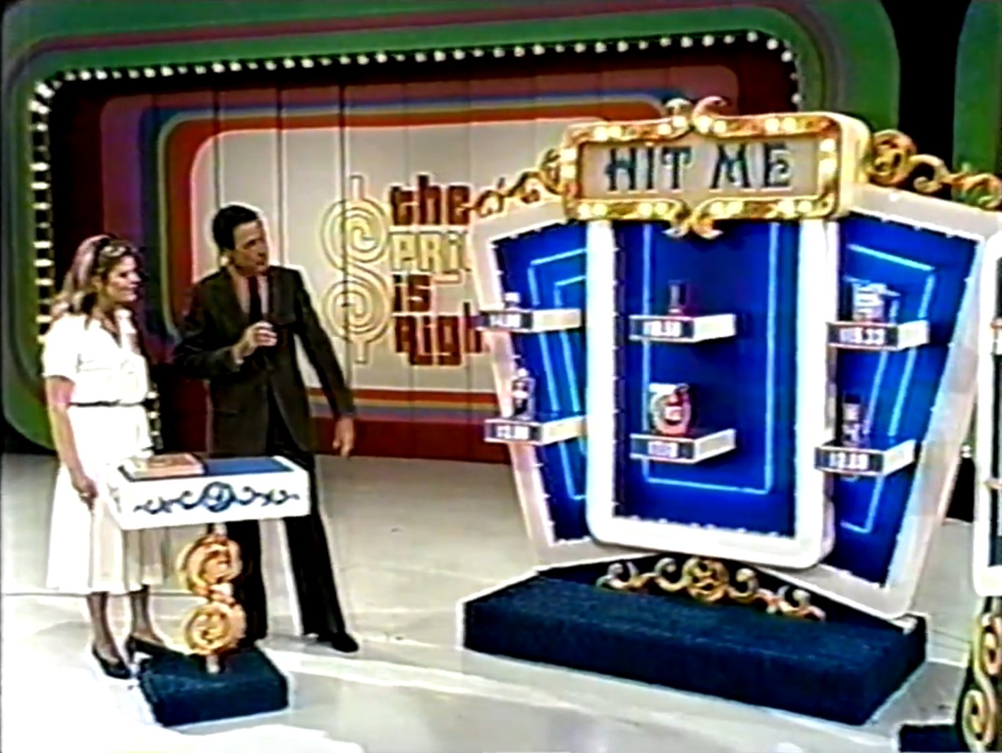 A television game show set featuring the 'Hit Me' game with two contestants. The set displays shelves holding grocery products with multiplied prices, while host Bob Barker engages with contestant Judith.