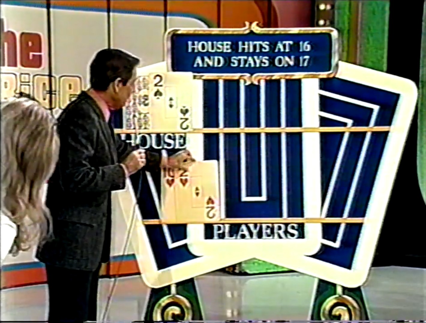 A game show host reveals the house's hand in a pricing game reminiscent of Blackjack, showing the house's two cards on a board with the player's beneath.