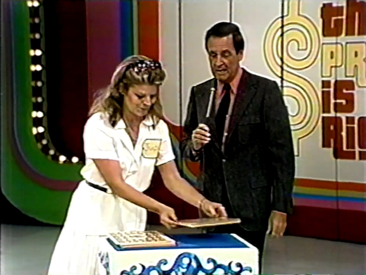 A woman with long hair and a white dress stands at a game table, cutting an oversized deck of cards while Bob Barker with a microphone speaks beside her. The colorful backdrop features the logo of 'The Price is Right'.
