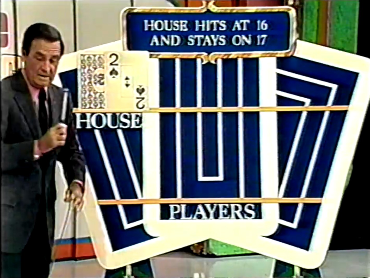 Game show host Bob Barker stands next to a large display board showing the house's hand in a Blackjack-inspired game. The board features visible cards, with a clear display indicating that the house hits on 16 and stays on 17.