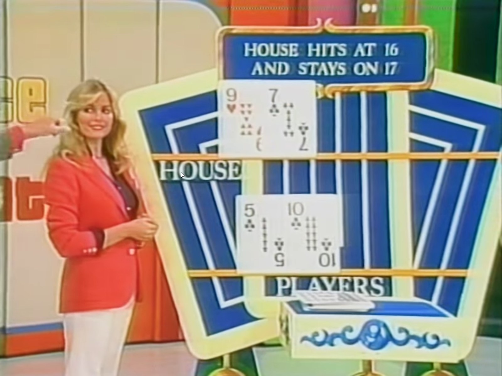 Game show host points to the scoreboard showing the house's hand with a 9 and 7, while a contestant stands beside them, smiling. The sign indicates that the house hits at 16 and stays on 17.