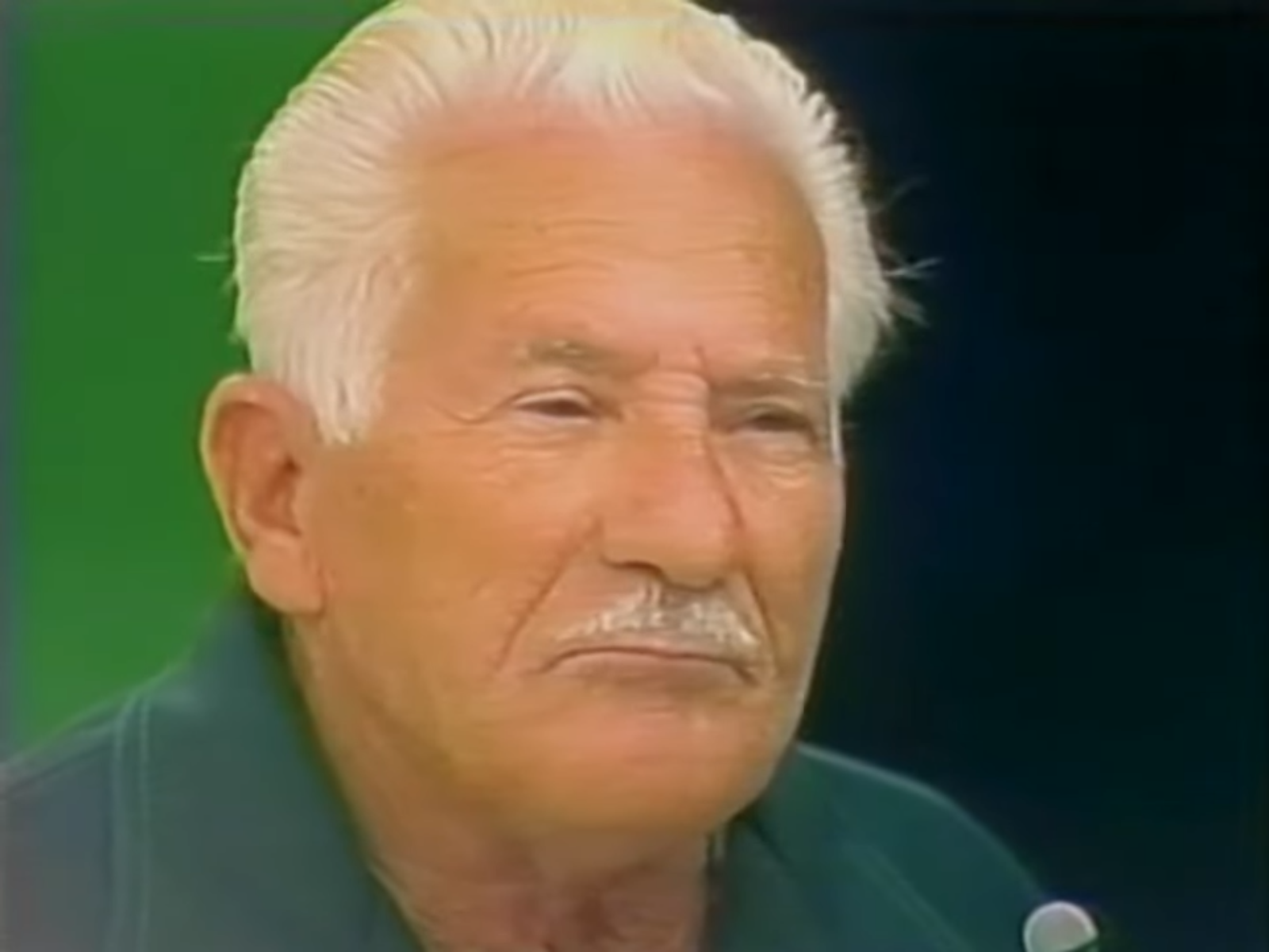 Close-up of an older man with white hair and a mustache, looking serious against a green background.