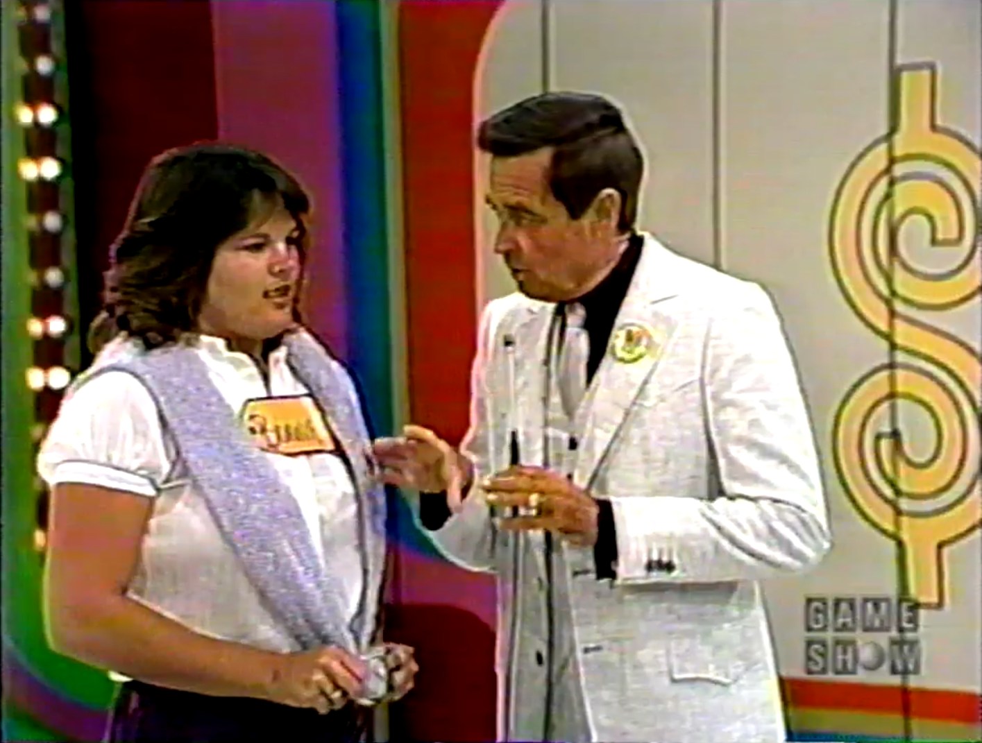 A game show host explaining the rules of 'Hit Me' to a contestant on stage, with bright multi-colored background.