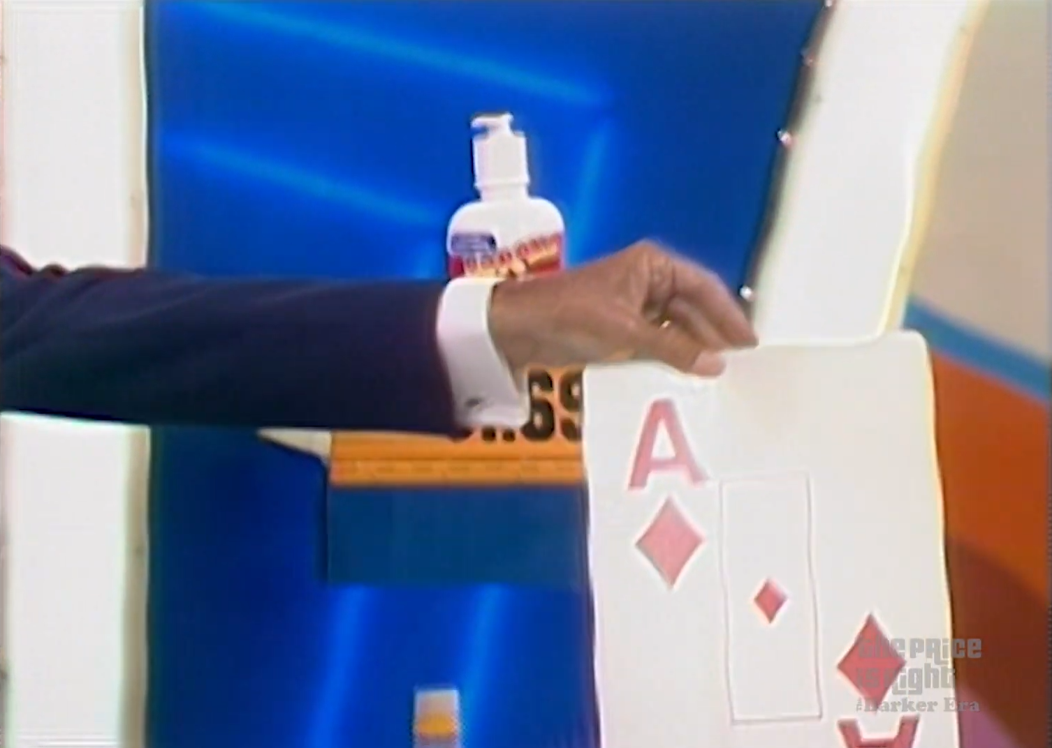 A hand holding an Ace of Diamonds card, displayed in a game show setting with a blue background and a bottle visible in the background.