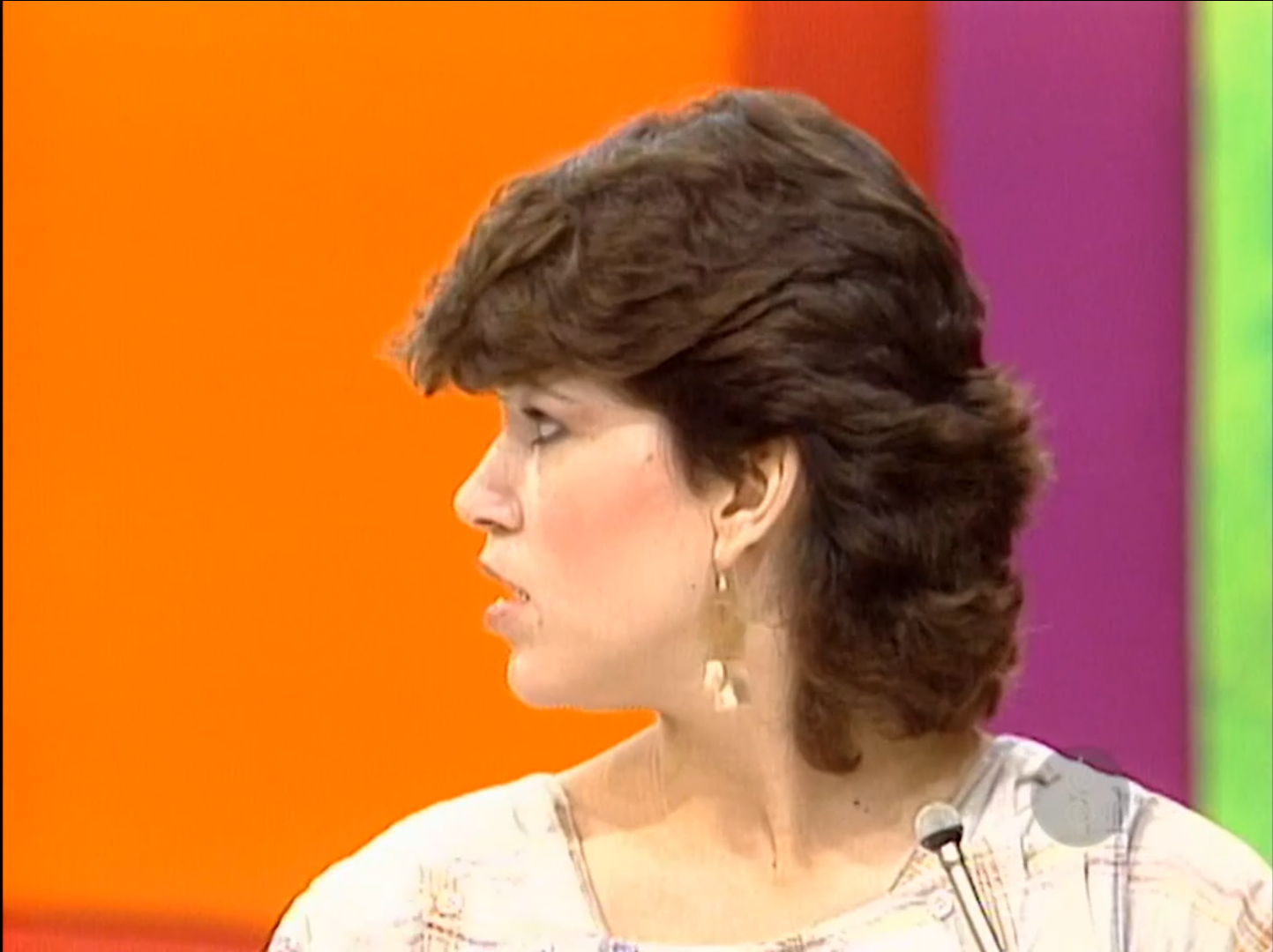 A woman with short hair facing to the side, wearing earrings and a light-colored patterned shirt, with a vibrant orange background.