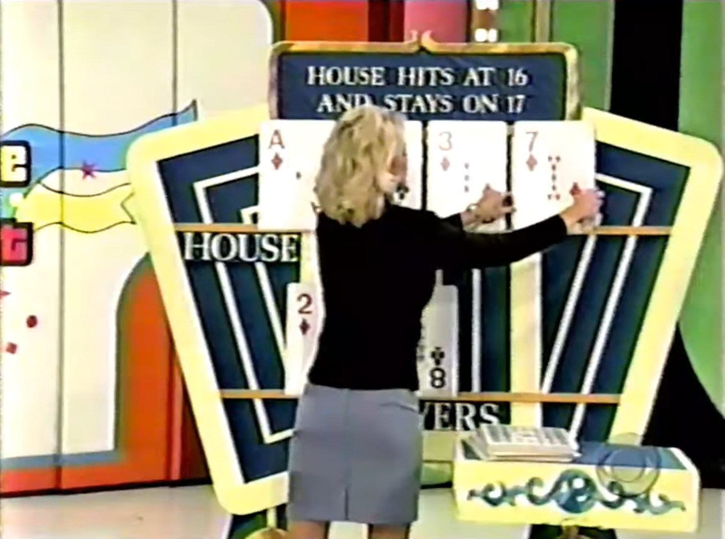 A model reveals cards for the house during the game 'Hit Me' on a pricing show, with the text 'HOUSE HITS AT 16 AND STAYS ON 17' visible.