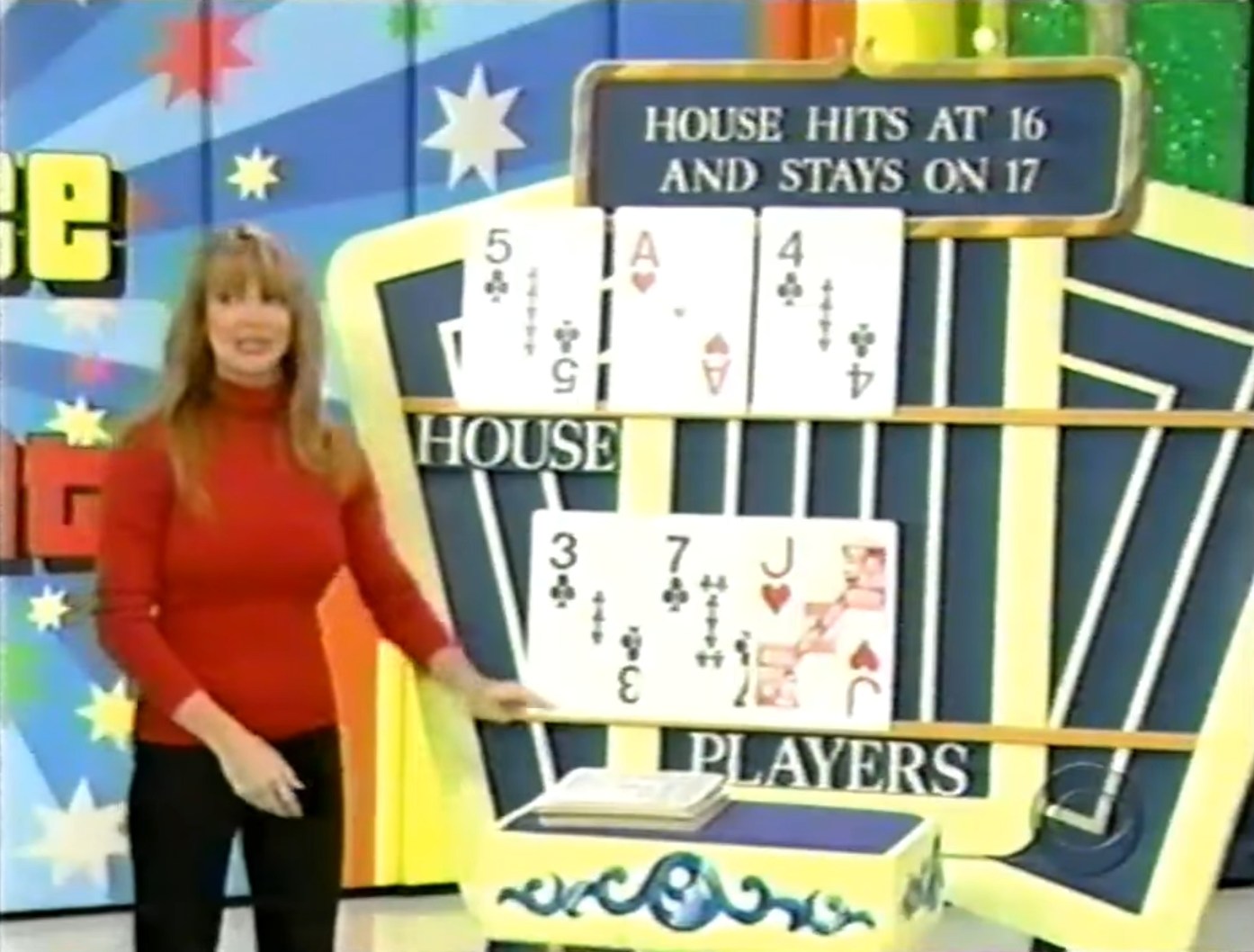Contestant presenting the game 'Hit Me' with displayed cards: House has a 5, Ace, and 4, while the Player has a 3, 7, and Jack, with a backdrop of colorful stars.