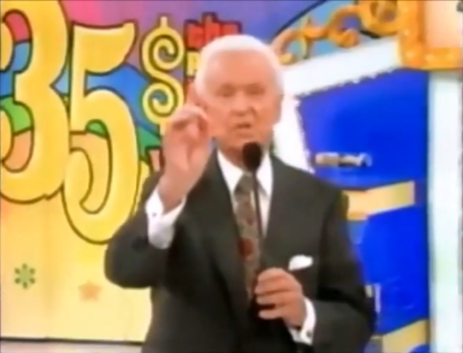 Bob Barker holds a microphone while gesturing towards a colorful 'Hit Me' game display on the 35th season of The Price is Right.
