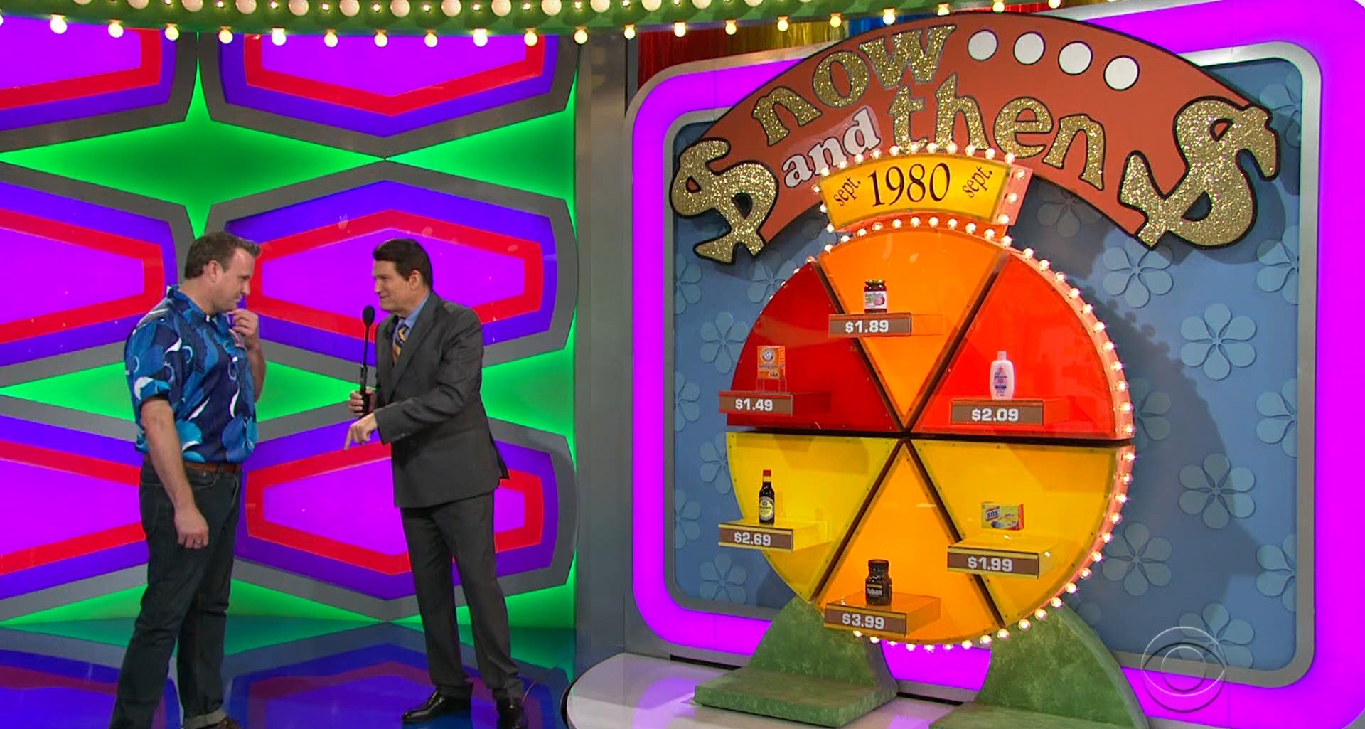 A game show contestant in a blue patterned shirt stands next to Drew Carey with black hair, discussing prices displayed on a colorful circular game board labeled 'Now and Then' with various grocery products and their prices from September 1980.