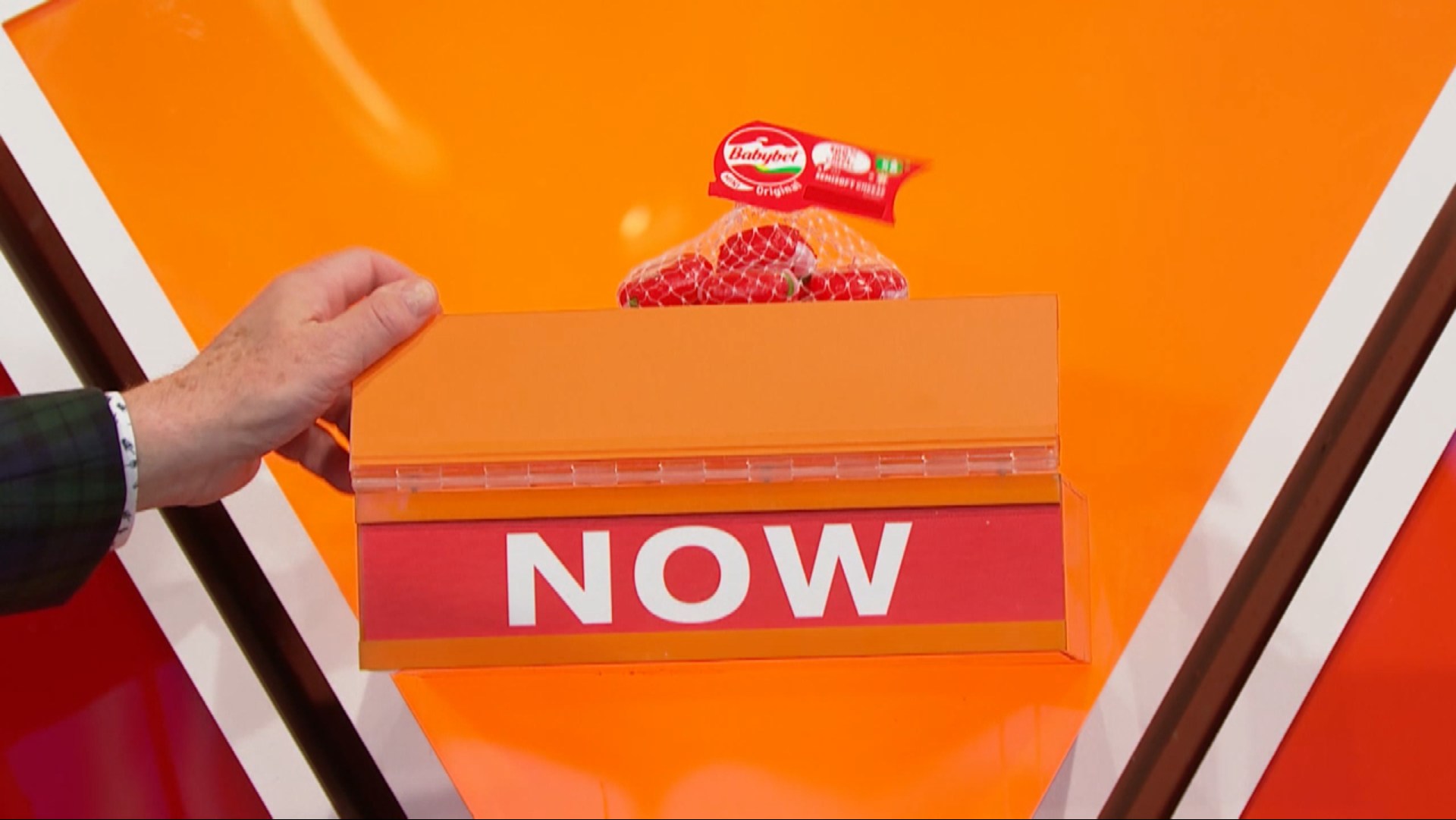 A hand holds an orange box displaying the word 'NOW' with a netted package of Babybel cheese on top against a bright orange background, proving "The Cheese is Now!"