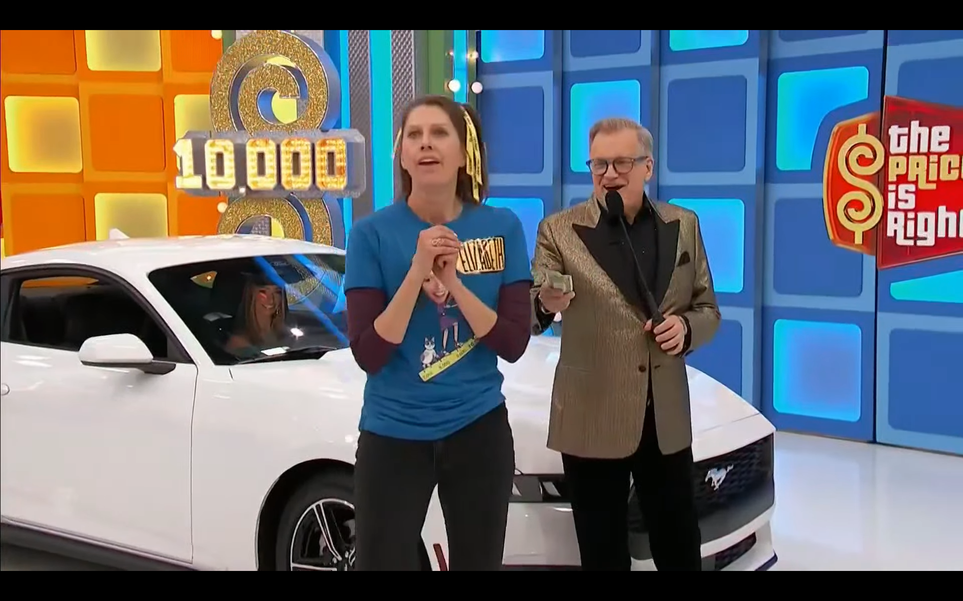 A contestant appears excited while standing next to a white car on the set of The Price is Right, with a host holding cash and a large display showing '10,000' in the background.