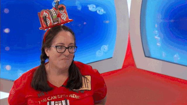 A contestant smiling while wearing a hat shaped like a stage with colorful decorations, standing in front of a brightly lit game show set.