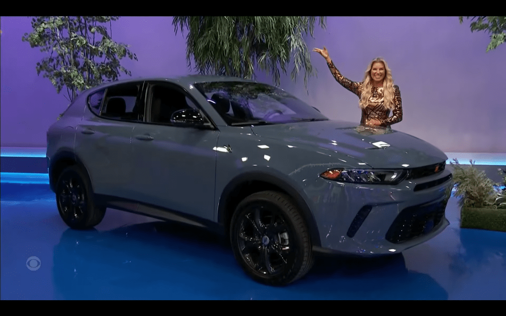 A model smiles and gestures next to a grey SUV, displayed on a brightly colored stage with plants in the background.