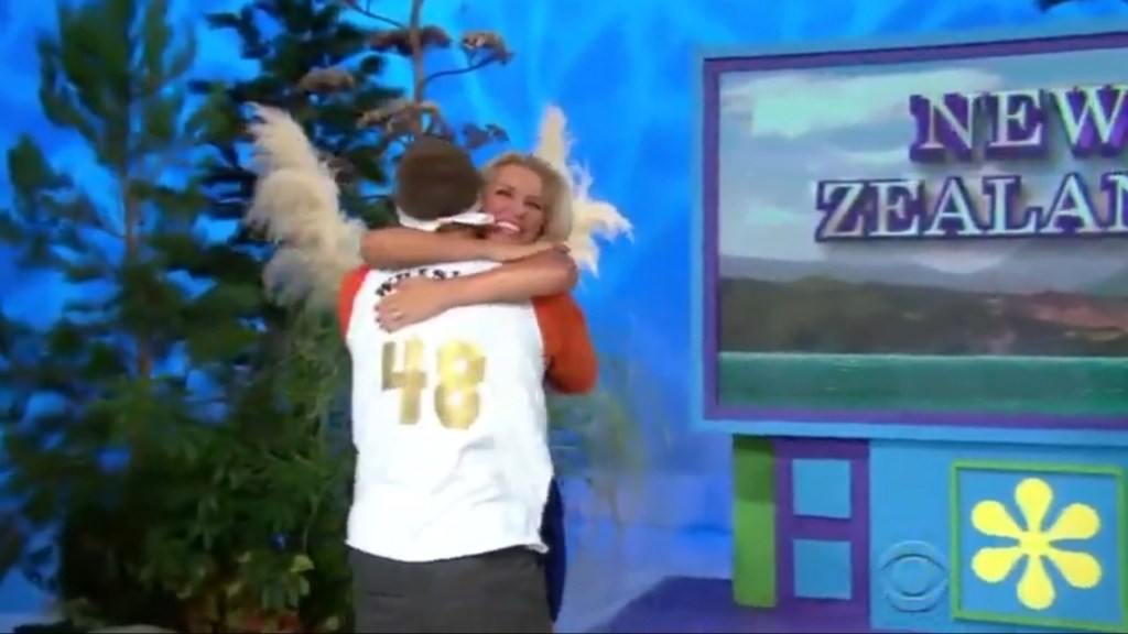 Contestant Steven hugs model Rachel Reynolds on The Price is Right after winning a trip to New Zealand, with vibrant set decorations in the background.