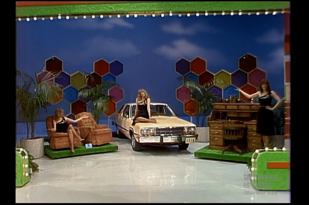 A game show stage featuring two models beside a pair of recliners and a wooden roll-top desk, with a car parked in the center. Colorful hexagonal background elements and decorative plants enhance the setting.