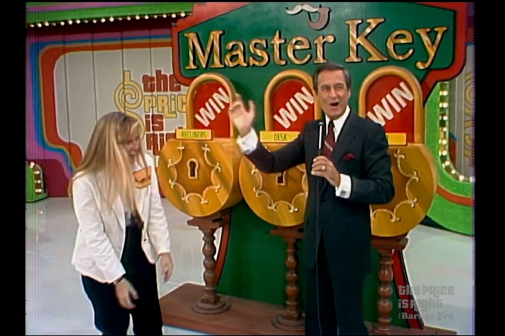 A contestant holding a prize key stands with a host beside the Master Key game display, featuring three locks labeled 'WIN' and 'Desk', with colorful decorations in the background.