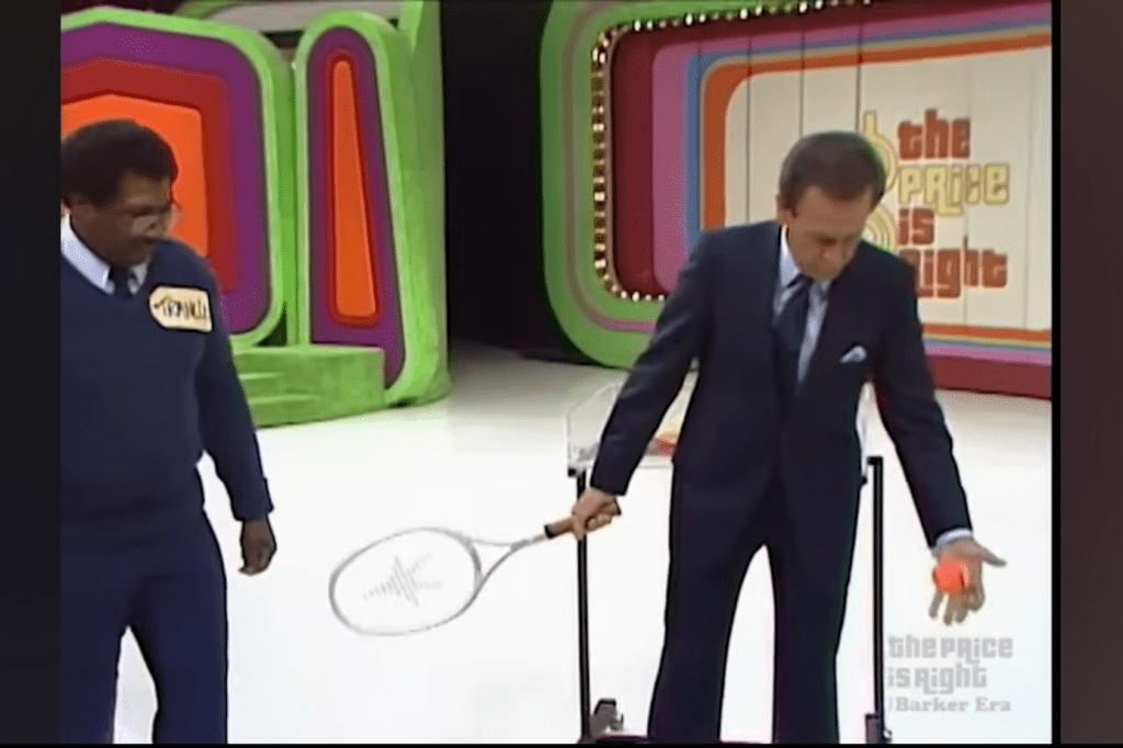 A contestant holds a tennis racket and an orange tennis ball while another person looks on, during a game on 'The Price is Right'. The colorful set features geometric shapes in the background.