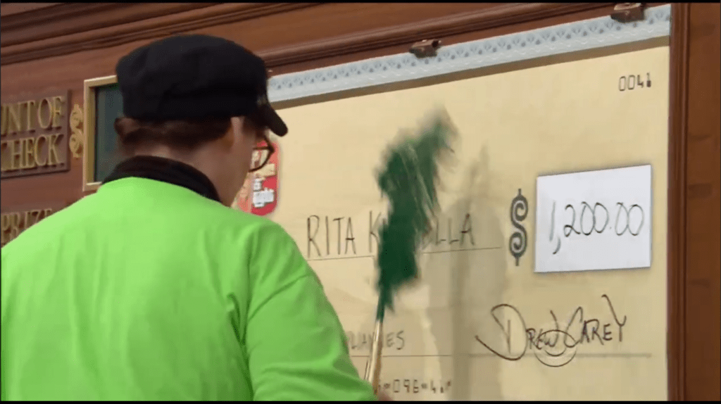 A contestant in a green shirt is seen filling out a large novelty check with the amount of ,200.00, while a host observes.