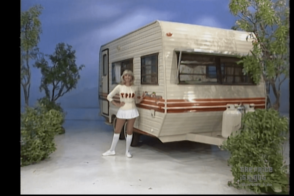 A model stands beside a camping trailer on a game show set, surrounded by greenery and a blue backdrop.
